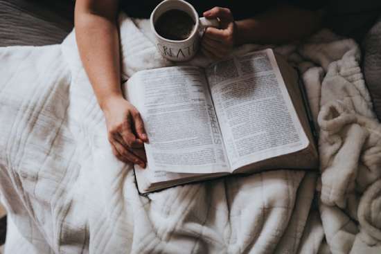 A person holding a mug of tea and reading the Bible while snuggled up in a blanket