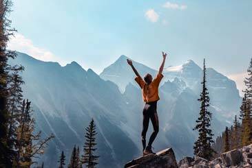 Person standing on a rock across a mountain range holding both hands up in the air beside trees under a clear blue sky - AAAF