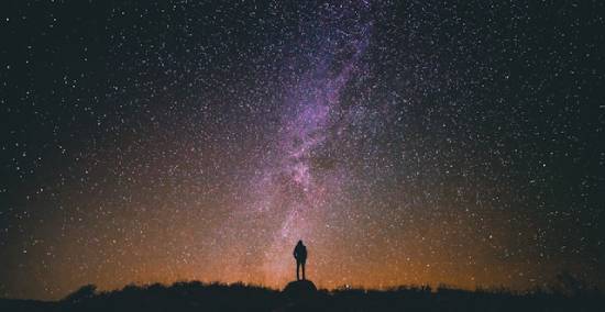 A person stands on a hill at night and gazes into a sky filled with stars