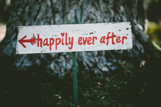Sign board of Happily ever after written with an arrow in red color on white board, fixed on ground near a tree - AAAF