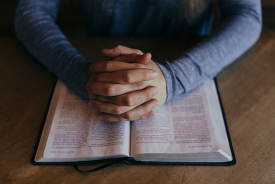 Man's hands praying on an open Bible as we prayerfully search the Scriptures diligently as our guide for testing prophets.