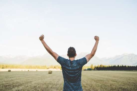 A man raises his arms over his head as he looks out over fields and mountains and enjoys the benefits of fresh air