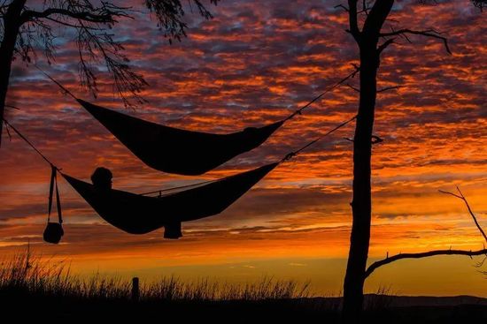 Person lying on one of the two hanging hammocks tied to trees under red sky at sunset. AAAF