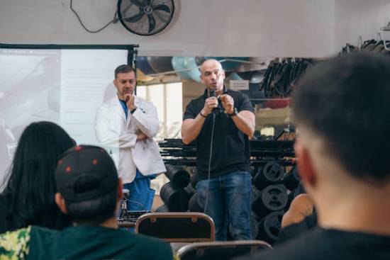 Man giving health seminar at a gym accompanied by a doctor A man speaks with a microphone to a small group; a doctor stands behind him.