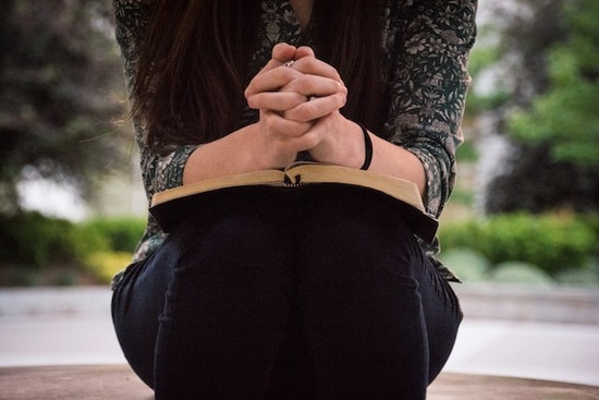 Woman sitting while praying with folded hands on an open Bible as we learn about various physical postures during prayer