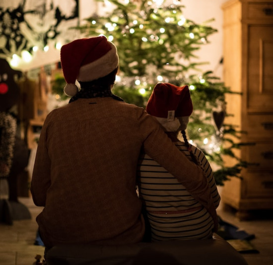 A parent and child wearing Santa hats and sitting by a Christmas tree