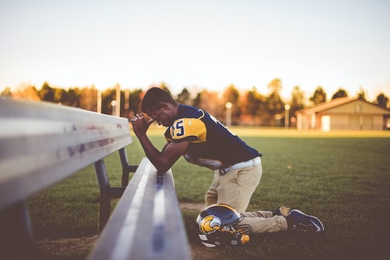 A football player kneeling in prayer at a bench