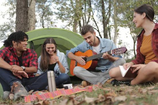 Four friends hanging out at a campsite while one of them plays a guitar. This illustrates the sense of fellowship and community Adventists may experience at camp meetings.
