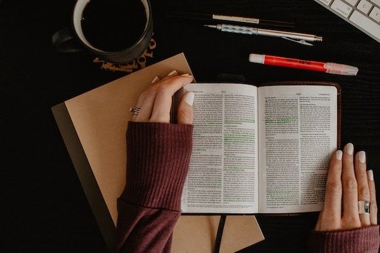 Hands holding a Bible open with pens and other study materials beside