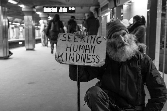 A homeless man with a cardboard sign that says, Seeking human kindness