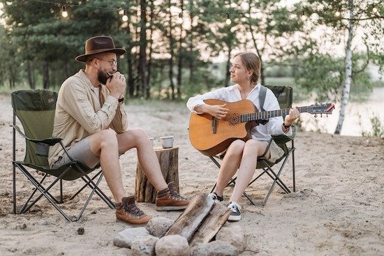 A man playing a harmonica and a woman playing a guitar by a campfire