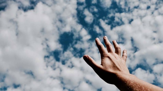 Hand stretched with open palm towards the sky filled with white clouds, reaching out to God in hope and faith