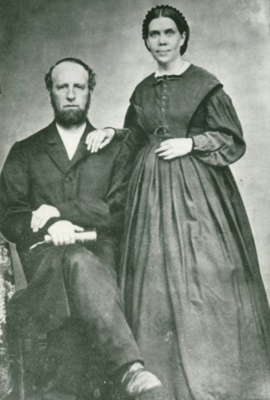 A black-and-white photograph shows James White sitting in a chair while his wife, Ellen White, stands by his side.