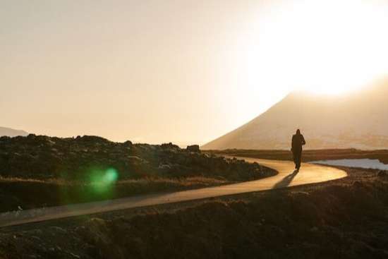 A man walking on a road with the sun setting behind a mountain ahead of him