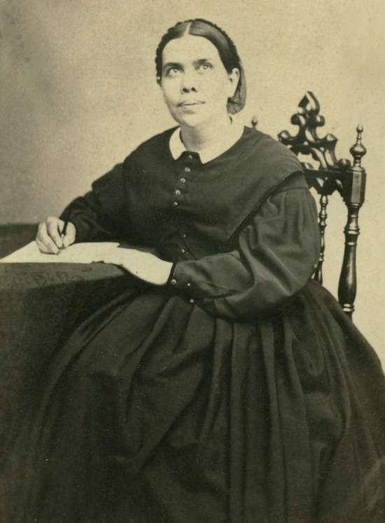 A black and white photograph of Ellen G. White sitting at a desk writing a manuscript.