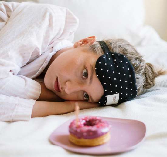 A girl laying in bed and resisting the temptation to eat a donut on a plate in front of her