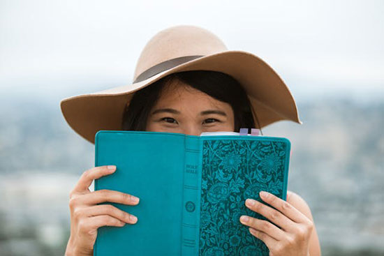Woman holding a Bible in front of her face a young woman with a hat on smiles with her eyes, an open Bible covering the bottom half of her face.