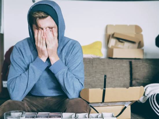 Sad Young Man Covering his Face with his Hands A young man in a hoody sits on a couch with his head in his hands, distraught about something.