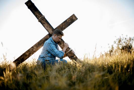 Man carrying a cross in a field A man carries a cross over his shoulder as he walks through a field, illustrating Jesus' teaching about taking up our crosses (Mark 8:34).