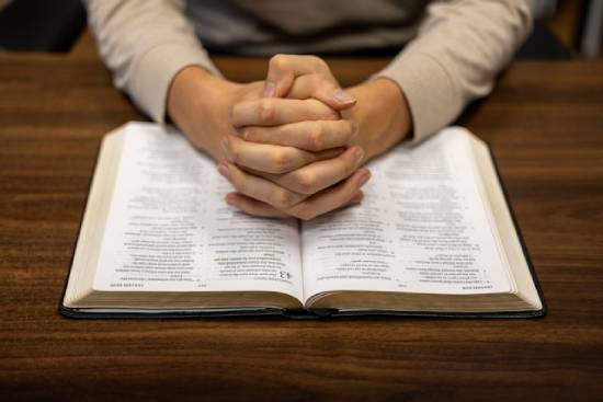 A person sitting at a table with their hands clasped on an open Bible A person clasps their hands together on top of an open Bible, signifying prayer