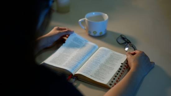 A woman reads her Bible with a mug and glasses nearby. This illustrates studying God's Word and taking it to heart.