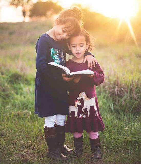 Kids Reading Bible An older sister embraces her younger sister as they stand outside in the sun and read the Bible