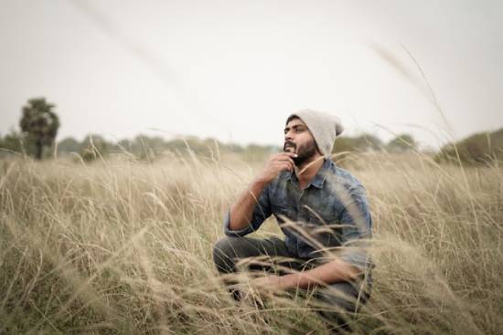Man in a field contemplating his relationship with God A young man of color squats in tall grass and contemplates something.