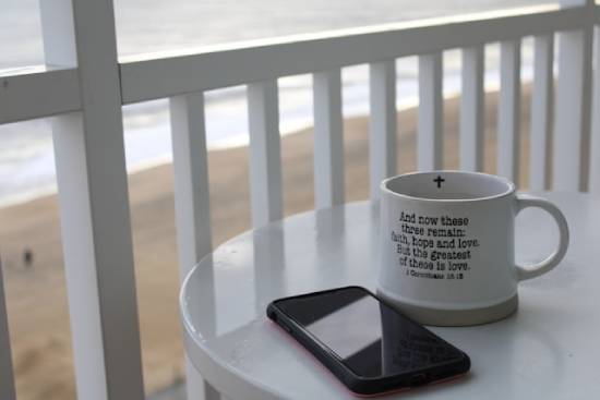 1 Corinthians 13:13 on a coffee mug A cellphone and mug with 1 Corinthians 13:13 written out on it sit on a beachfront table.
